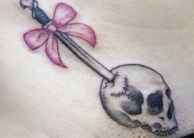 Fine line tattoo of a skull with a dagger and pink bow, in the size of a quarter.